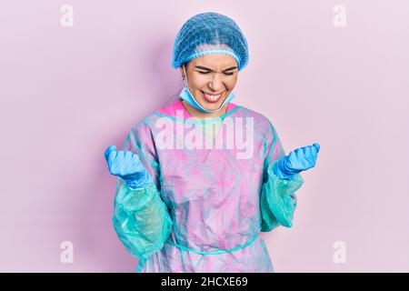 Giovane donna bruna indossando chirurgo uniforme e maschera medica molto felice ed eccitato facendo il gesto del vincitore con le braccia sollevate, sorridenti e urlanti per Foto Stock