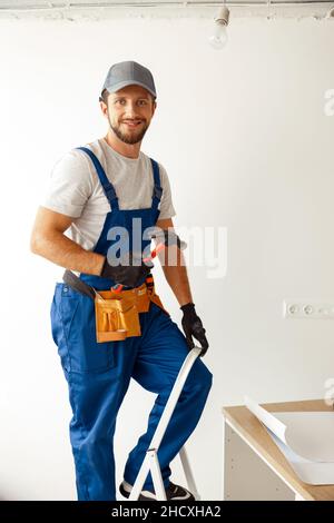 Elettricista allegro in uniforme sorridendo alla macchina fotografica, tenendo il martello mentre si installa la luce di montaggio nel nuovo appartamento Foto Stock