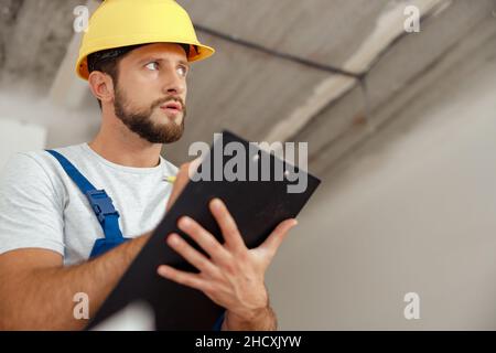 Il riparatore pensivo in abiti da lavoro e hardhat tenendo appunti e scrivere dettagli di un ordine mentre si trova in piedi al coperto durante i lavori di ristrutturazione Foto Stock