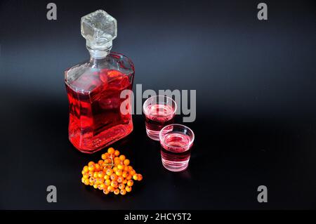 Liquore Rowan, alcol fatto in casa in due bicchieri e una bottiglia accanto a grappoli di bacche mature su sfondo nero. Primo piano. Foto Stock