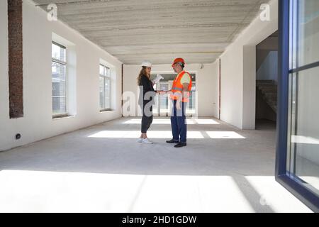Persona che indossa il cappuccio di sicurezza e il gilet arancione che scuote le mani con un ingegnere femminile in tuta nera Foto Stock