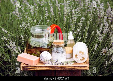 Vari prodotti spa lavanda su vassoio di legno in un campo di lavanda in fiore nella giornata estiva soleggiata. Caraffa di vetro riempita con una bevanda calda al tè alla lavanda. Foto Stock