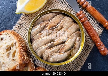 Spratto affumicato con olio in lattina aperta, pane intero di grano, limone su sfondo nero. Vista dall'alto, piatto. Foto Stock