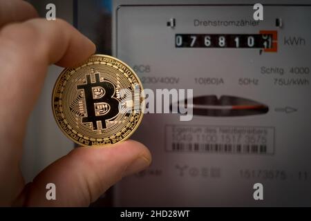 una mano che tiene una moneta di bitcoin davanti ad un contatore trifase Foto Stock