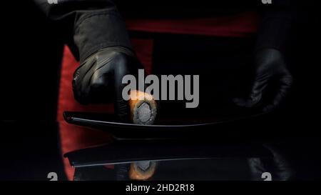 Lo chef del sushi mette il sushi roll sul piatto. Primo piano chef in grembiule nero mani in guanti mette un rotolo di sushi sul piatto e lo porta via. Foto Stock