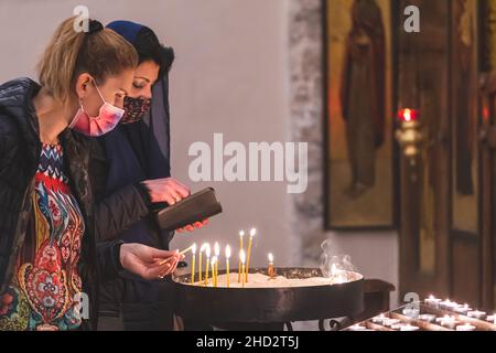 Due belle ragazze caucasiche o donne che indossano maschere e illuminazione candele votive in una chiesa Foto Stock