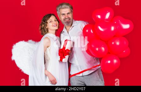 Buon giorno di San Valentino. Coppia sorridente con palloncini a forma di cuore rosso e regalo. Coppia Angeli Cupido Foto Stock