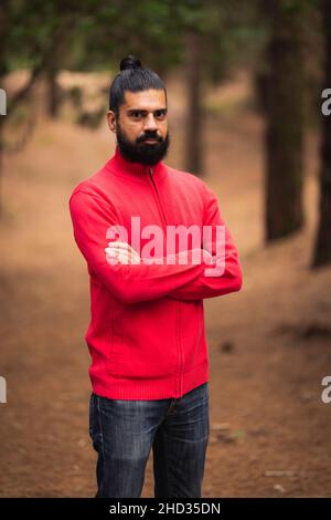 Colpo verticale di un uomo ispanico barbuto che indossa un maglione rosso nella foresta Foto Stock