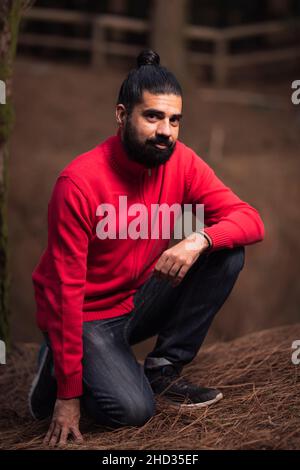 Colpo verticale di un uomo ispanico inginocchiato che indossa un maglione rosso nella foresta Foto Stock