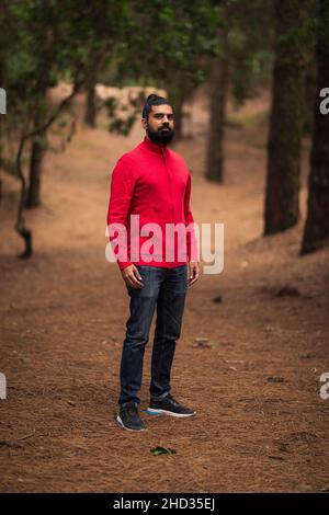 Colpo verticale di un uomo ispanico barbuto che indossa un maglione rosso nella foresta Foto Stock