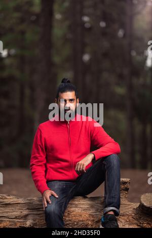 Un colpo verticale di un uomo ispanico barbuto che indossa un maglione rosso nella foresta Foto Stock