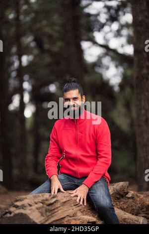 Colpo verticale di un uomo ispanico sorridente con una barba seduta su un tronco d'albero nella foresta Foto Stock