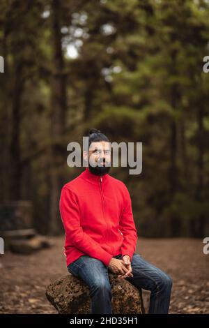 Colpo verticale di un uomo ispanico barbuto seduto su un ceppo di albero nella foresta Foto Stock