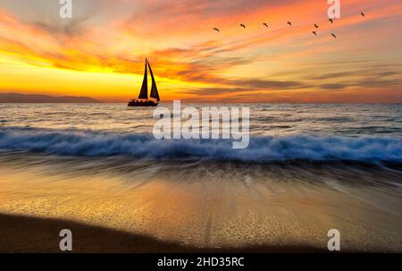 Una barca a vela sta navigando lungo l'oceano con un gregge di uccelli che volano sopra UN cielo colorato di tramonto Foto Stock