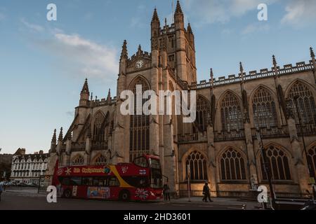 Un autobus turistico rosso a due piani passa davanti alle finestre gotiche e ai contrafforti dell'Abbazia di Bath nella storica città di Bath, Somerset, Inghilterra, Regno Unito Foto Stock