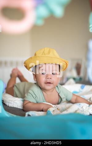 Adorabile 2 mesi di razza mista neonato bambino ragazzo indossare un cappello e sdraiato sul pancino sul letto sollevare testa e giocare circondato da giocattoli Foto Stock