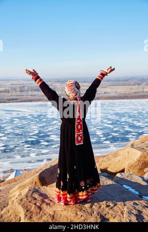 Vista posteriore di donna anonima in abito tradizionale e cappello che solleva le braccia e godere della luce del sole con gli occhi chiusi, mentre in piedi sulla scogliera contro congelato Foto Stock