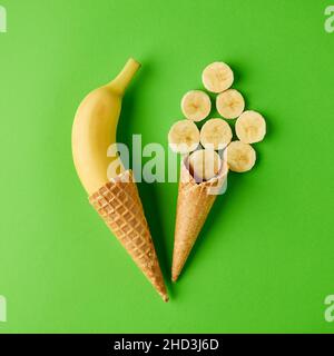 Coni gelato riempiti con una banana intera e fette di banana su sfondo verde chiaro. Concetto minimo di cibo per la vita morta creativa. Foto Stock