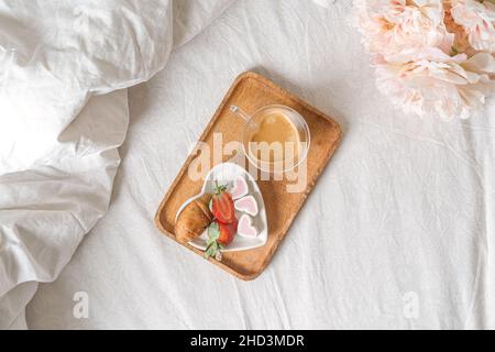 Colazione per San Valentino. Piatto bianco a forma di cuore con fragole fresche, tazza di caffè, regalo e fiori con regalo a letto. Vita morta Foto Stock