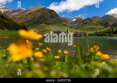 Il Santuario di Núria (Santuari de la Mare de Déu de Núria) con il suo lago e palude-marigold o kingCup (Caltha palustris) fiori sfocati Foto Stock