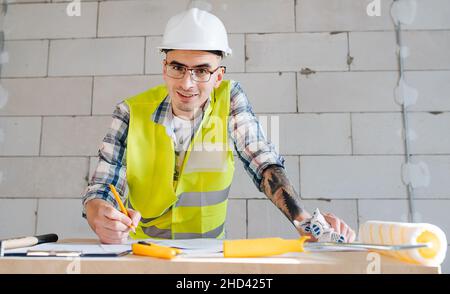 Gentile e simpatico ingegnere edile in casco bianco che si disegna su un progetto su un tavolo improvvisato. Su un muro di mattoni grigio ruvido. Guardando Foto Stock