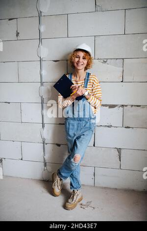 Sorridente ingegnere di costruzione femminile in un casco bianco che tiene una clipboard, appoggiata su una parete di mattoni grigio ruvida. Foto Stock