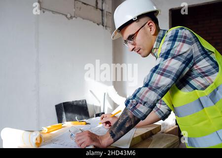 Vista laterale di un pensieroso ingegnere edile in casco bianco che si disegna su un piano su un tavolo improvvisato. All'interno di un edificio incompiuto. Foto Stock