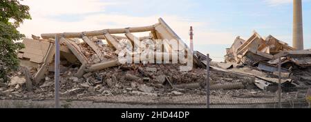 Costruzione industriale di calcestruzzo dietro una recinzione distrutta da sciopero. Scenario di disastro pieno di detriti, polvere e edifici schiantati con il sole crescente e blu s Foto Stock