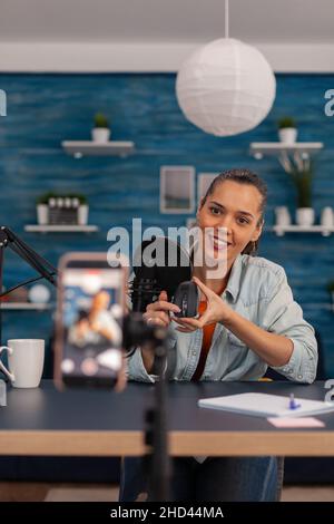 Creatore di contenuti che riprende la revisione del mouse con il telefono cellulare. Influencer donna in studio domestico con microfono e test dei prodotti di registrazione smartphone. Podcast vlog online per i social media. Foto Stock