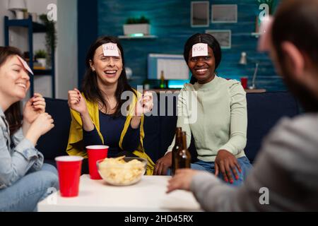 Un gruppo di amici che ride giocando a un gioco di gusess usando le note adesive che si divertono a passare il tempo insieme durante la festa di wekeend a casa. Persone multietniche che bevono birra indovinando la risposta corretta Foto Stock