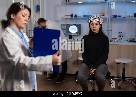 Neurologo specialista che tiene appunti con informazioni sulla tomografia durante l'esperimento neuroscientifico in laboratorio moderno. Medico ricercatore che monitora l'attività cerebrale del paziente con la cuffia eeg Foto Stock