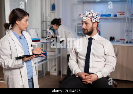 Neurologo donna medico tipizzazione sintomi di malattia neurologica su tablet computer discutere trattamento medico con il paziente. Ingegnere ricercatore che monitora l'attività cerebrale dell'uomo con cuffie eeg Foto Stock