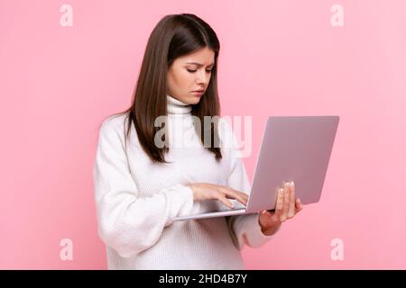 La vista laterale ritratto della ragazza pensiva funziona in linea, guarda il display del laptop con espressione premurosa, indossando il maglione casual bianco di stile. Studio interno girato isolato su sfondo rosa. Foto Stock