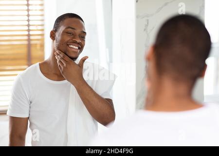 Cura della pelle maschile. Bel ragazzo nero che guarda nello specchio in bagno, toccando la barba Foto Stock