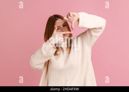 Ritratto di donna bionda che imita per scattare foto, guardando attraverso la cornice delle dita con vista attenta, messa a fuoco e ritaglio immagine, indossando maglione bianco. Studio interno girato isolato su sfondo rosa. Foto Stock