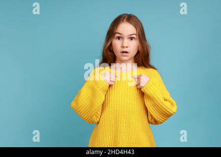 Ritratto di bambina guarda la macchina fotografica con il volto scioccato, si punta, essendo orgoglioso e sorpreso, indossando giallo casual stile maglione. Studio interno girato isolato su sfondo blu. Foto Stock