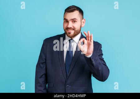 Va tutto bene. Uomo sorridente che indossa un abito di stile ufficiale, guardando una fotocamera nat, mostrando il segno ok, esprimendo emozioni positive. Studio interno girato isolato su sfondo blu. Foto Stock