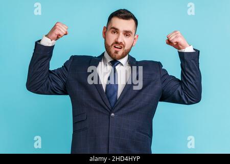 Sono forte. Ritratto di uomo d'affari motivato auto-sicuro in vestito di stile ufficiale osservando le braccia assertive e sollevando per mostrare bicipiti, ritenendo potente. Studio interno girato isolato su sfondo blu. Foto Stock