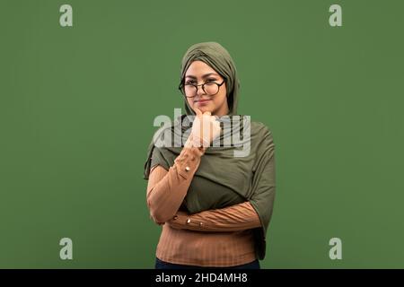 Donna araba pensiva in occhiali che toccano il mento e pensa alla domanda, in piedi su sfondo verde scuro, spazio copia Foto Stock