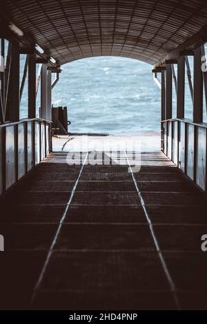 A vertical view of a descending boarding gate, a covered corridor with plenty of beams, on the quay for passengers of a boat or a ship, with river wat Foto Stock