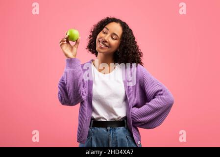 Ritratto di allegro giovane donna nera con mela verde matura e sorridente su sfondo rosa studio Foto Stock