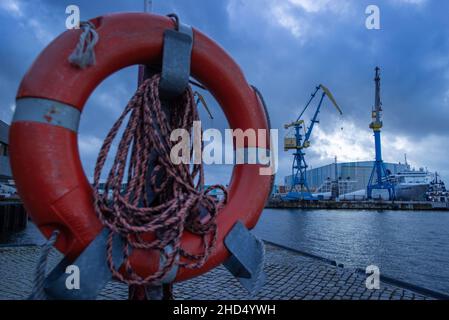 Wismar, Germania. 03rd Jan 2022. Un conservatore di vita si trova di sera nel porto della città di fronte al sito MV Werften con gru, una ex nave residenziale per i lavoratori dei cantieri navali e la grande sala di costruzione navale. Lo stato di Mecklenburg-Vorpommern non deve pagare per il momento un prestito di aiuto concordato di 78 milioni di euro (88 milioni di dollari USA) al proprietario di MV Werften Genting Hong Kong. La crisi a MV Werften, che attualmente conta circa 2.000 dipendenti e attività a Stralsund, Rostock e Wismar, continua a venire in testa. Credit: Jens Büttner/dp/dpa/Alamy Live News Foto Stock