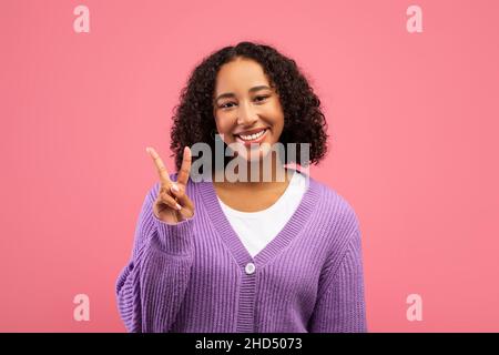 Umore positivo. Ritratto di gioiosa giovane donna nera che mostra il gesto di vittoria su sfondo rosa studio Foto Stock