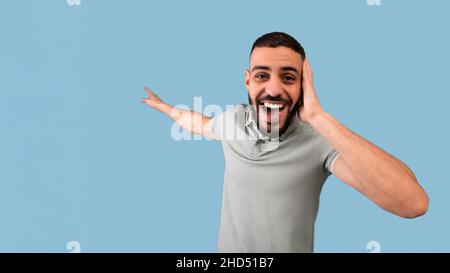 Shocked il tipo arabo che afferra la sua testa e punta allo spazio vuoto con la bocca aperta, offrendo gli sconti enormi, sfondo blu Foto Stock