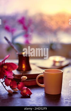 Caffè orientale cotto in tradizionale pentola di rame turco su vecchi libri. Tazza in ceramica in primo piano. Inverno con vecchi libri. Finestra del tramonto Foto Stock