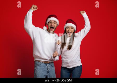 Coppia overjoyed in Santa Hats che celebra emotivamente il successo con Raised Fists Foto Stock