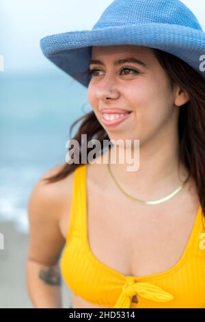 Ritratto di giovane donna in spiaggia in cappello blu e costume da bagno giallo Foto Stock