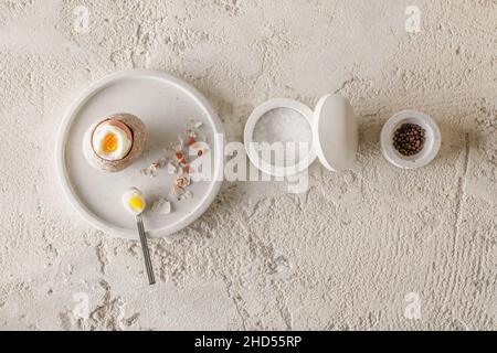 Un uovo sodo in una tazza di gres con guscio di uovo pezzi rotti in un vassoio di cemento, scaglie di sale e pepe in ciotole di cemento bianco su cl ruvido testurizzato Foto Stock