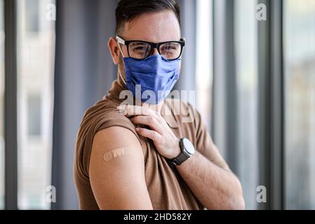 Giovane uomo adulto che mostra puntando al bendaggio sulla spalla del suo braccio indossare maschera protettiva fatto con la vaccinazione. Il maschio è appena stato vaccinato contro Covid1 Foto Stock