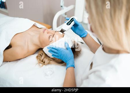 Cosmetologist in guanti blu fa ecografia pulizia del viso di una donna. Procedura di scrabbing a ultrasuoni Foto Stock